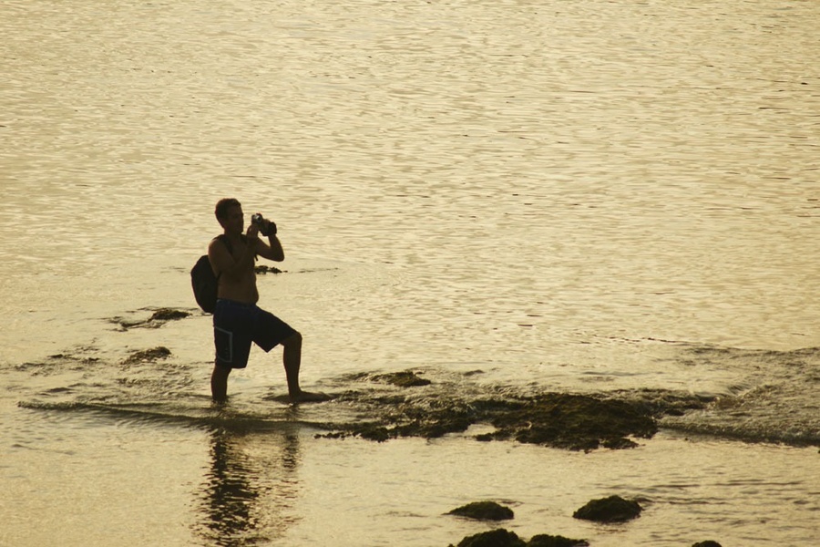 un hombre sin camisa está parado en la orilla del océano tomando una foto