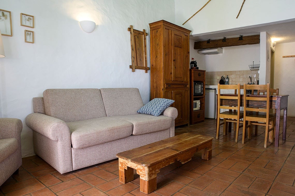 Sala de estar aconchegante na Aldeia da Pedralva com sofá, mesa de centro rústica e cozinha.