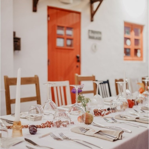 Uma mesa de jantar elegantemente posta com talheres, velas e frutas estende-se em frente a uma casa branca com uma vibrante porta e janela laranja.