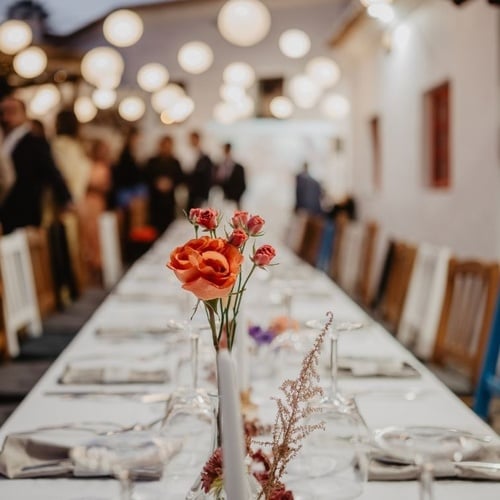 Uma longa mesa de jantar está preparada com arranjos florais e velas em primeiro plano, enquanto pessoas e luzes penduradas aparecem desfocadas ao fundo, criando um ambiente aconchegante.