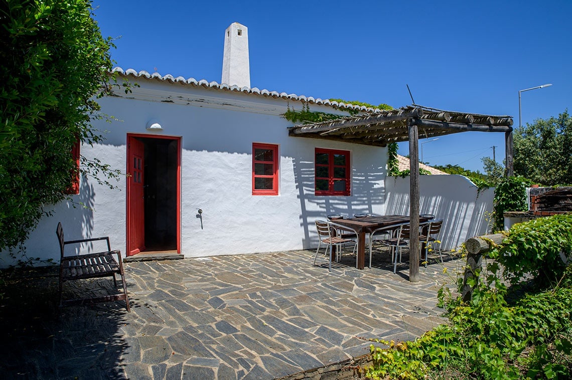Pátio rústico com mesa e cadeiras, porta vermelha e chaminé em Aldeia da Pedralva.