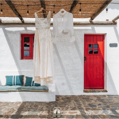 Uma fachada de casa branca com uma porta e janela vermelhas, um vestido e uma blusa brancos pendurados, um banco com almofadas azuis e um chão de pedra.