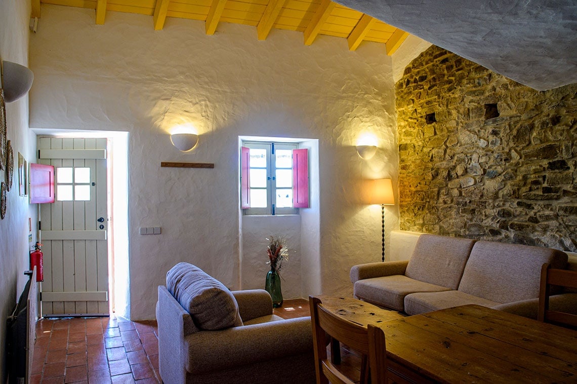 Sala de estar rústica na Aldeia da Pedralva com sofás, janela e porta tradicional.