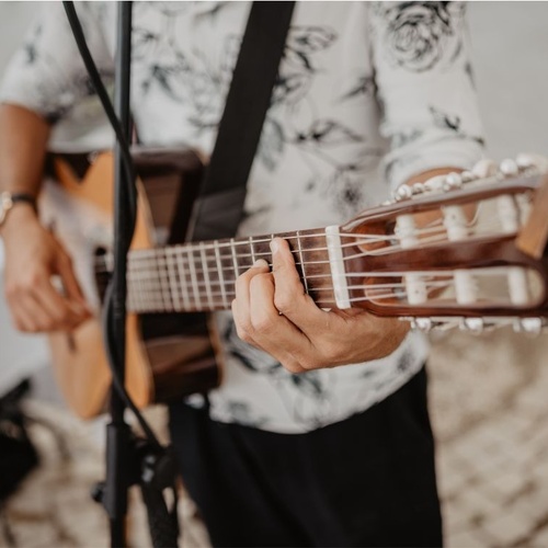 Uma pessoa com uma camisa estampada toca violão acústico, com as mãos no braço do instrumento e a cabeça do violão em foco.
