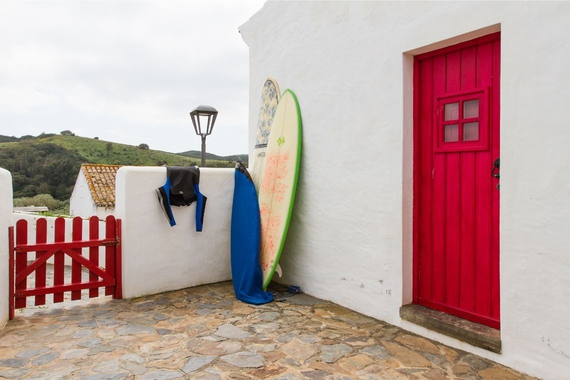 Una fachada blanca con una puerta y una valla rojas vibrantes, tablas de surf y un traje de neopreno colgando, sobre un suelo de piedra y con colinas verdes al fondo.