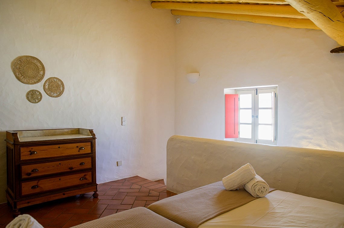 Quarto rústico na Aldeia da Pedralva com vista para a janela, dresser de madeira e camas arrumadas.