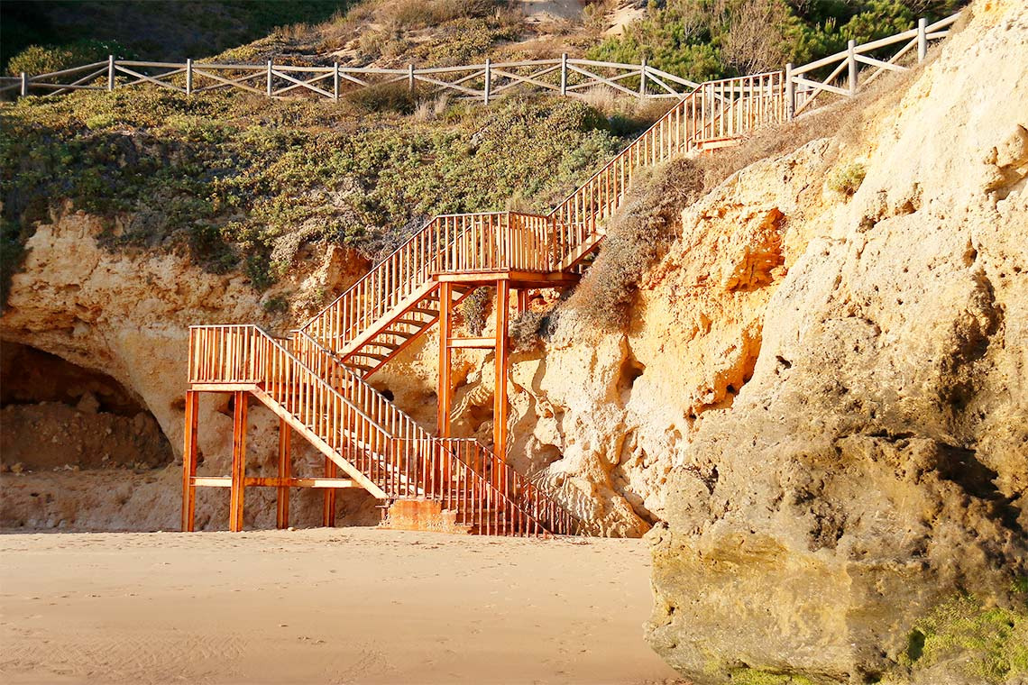 Wooden stairs lead up a sandy cliff to a Aldeia da Pedralva path and vegetated hillside.