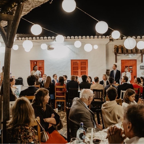 Uma receção noturna é iluminada por lanternas de papel brancas, com convidados sentados à mesa e oradores em destaque.