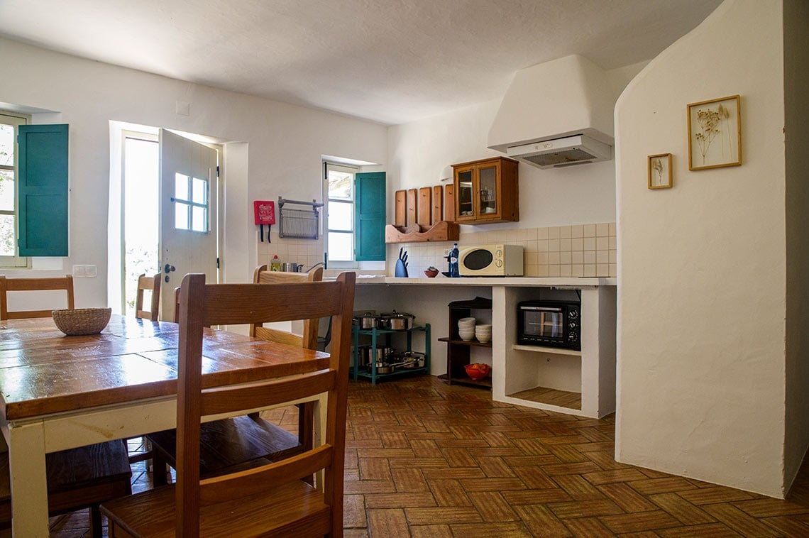 Cozinha rústica da Aldeia da Pedralva com mesa de madeira, cadeiras e eletrodomésticos.