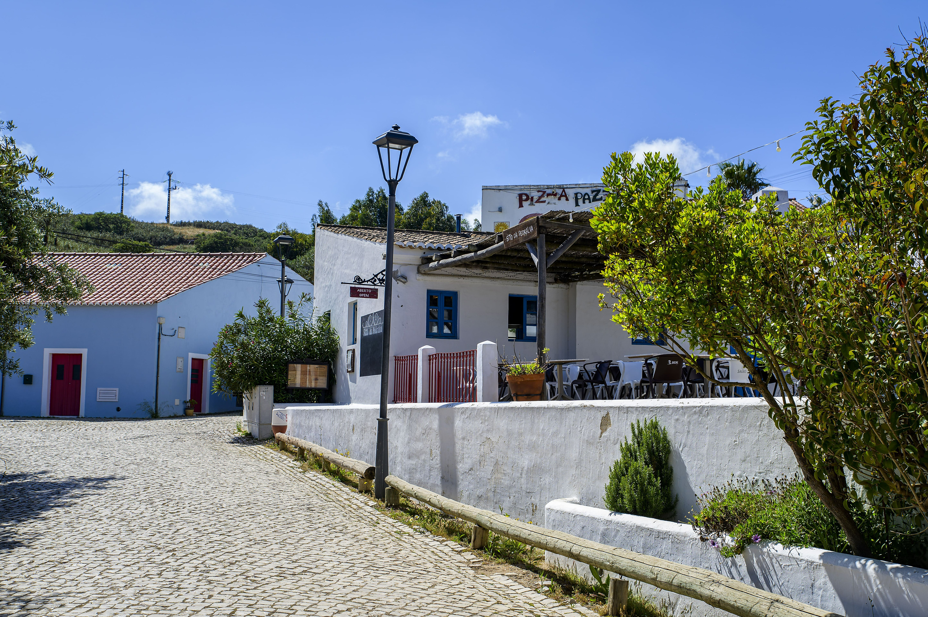 Fachada de restaurante 'Pizza Paz' com mesas ao ar livre e rua de paralelepípedos. Aldeia da Pedralva