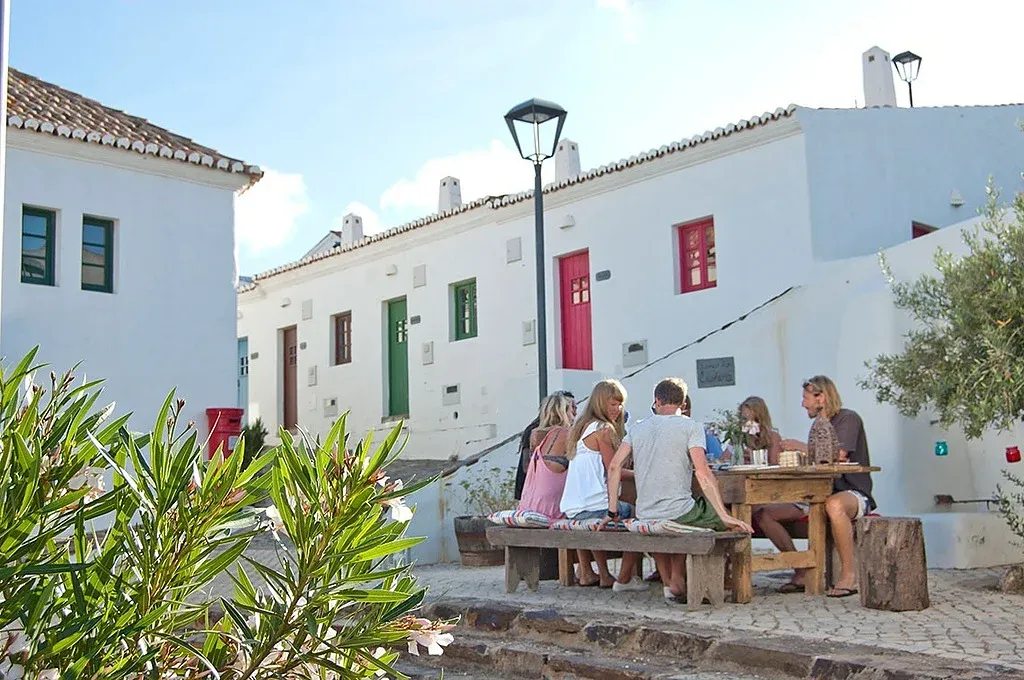 Amigos relaxam em mesas ao ar livre com a arquitetura branca típica deAldeia da Pedralva.