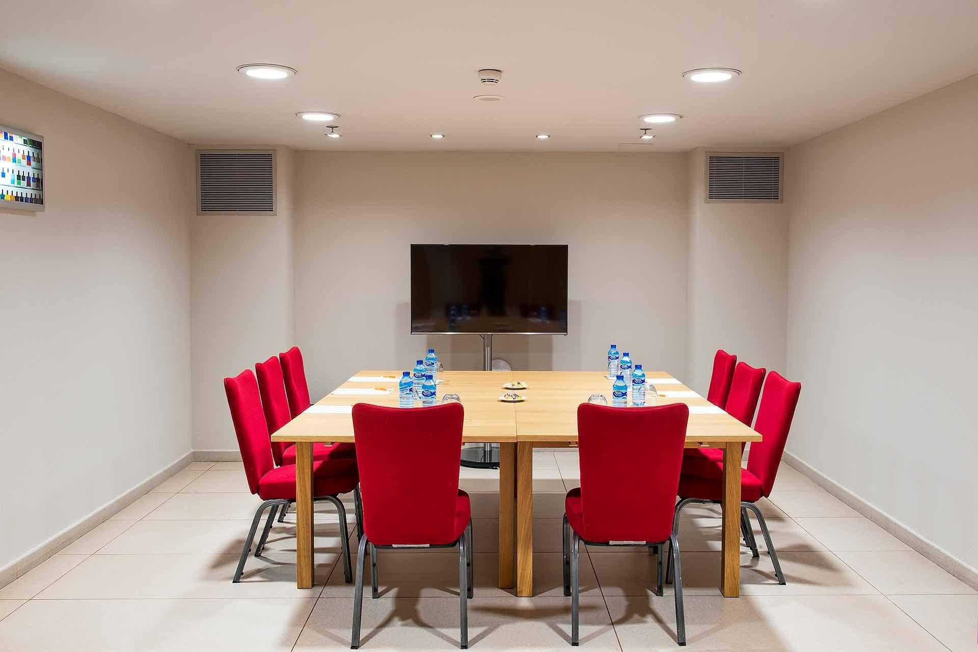Alexandre Corporativa meeting room with large wooden table, red chairs, TV, and water bottles.