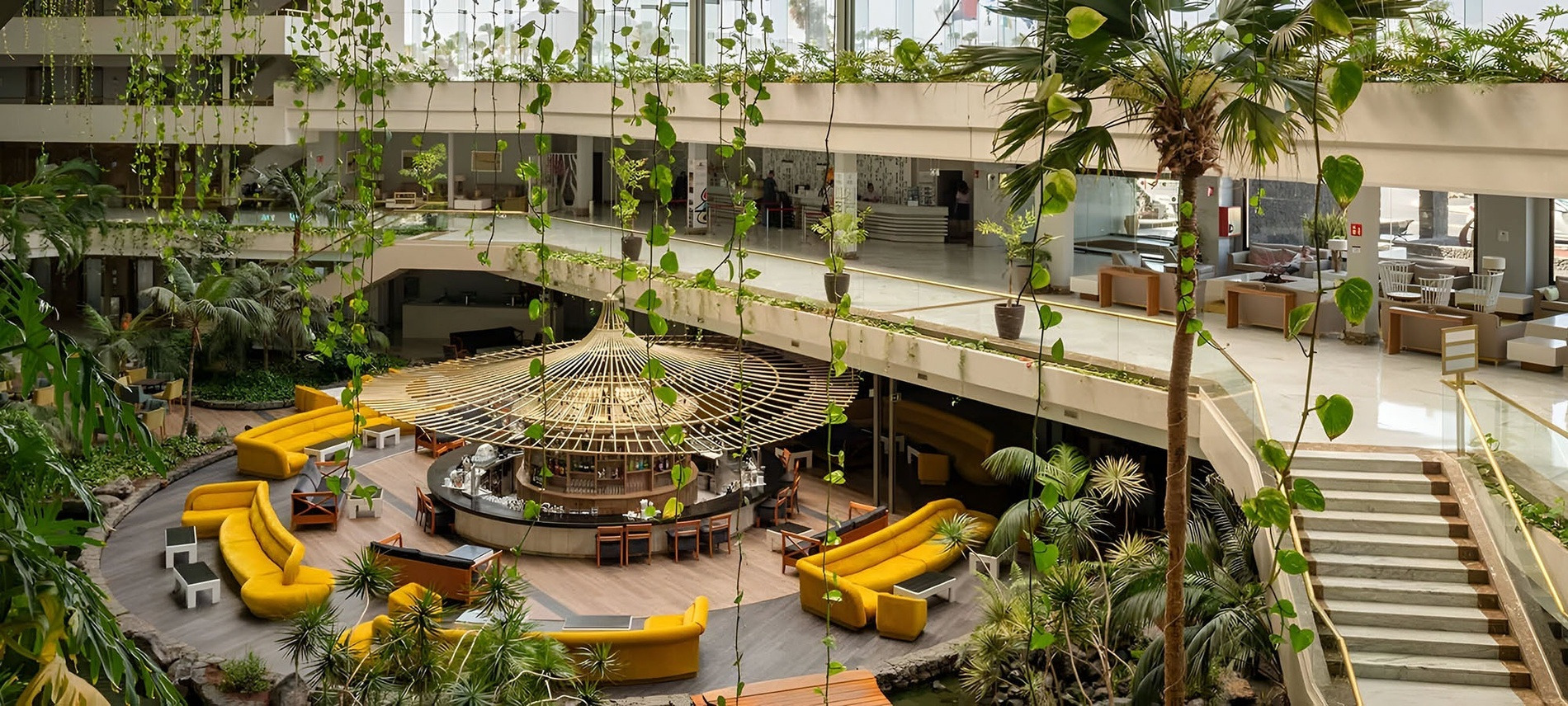 Lobby tropical de l'Hôtel Alexandre Corporativa avec bar central, palmiers et canapés jaunes.