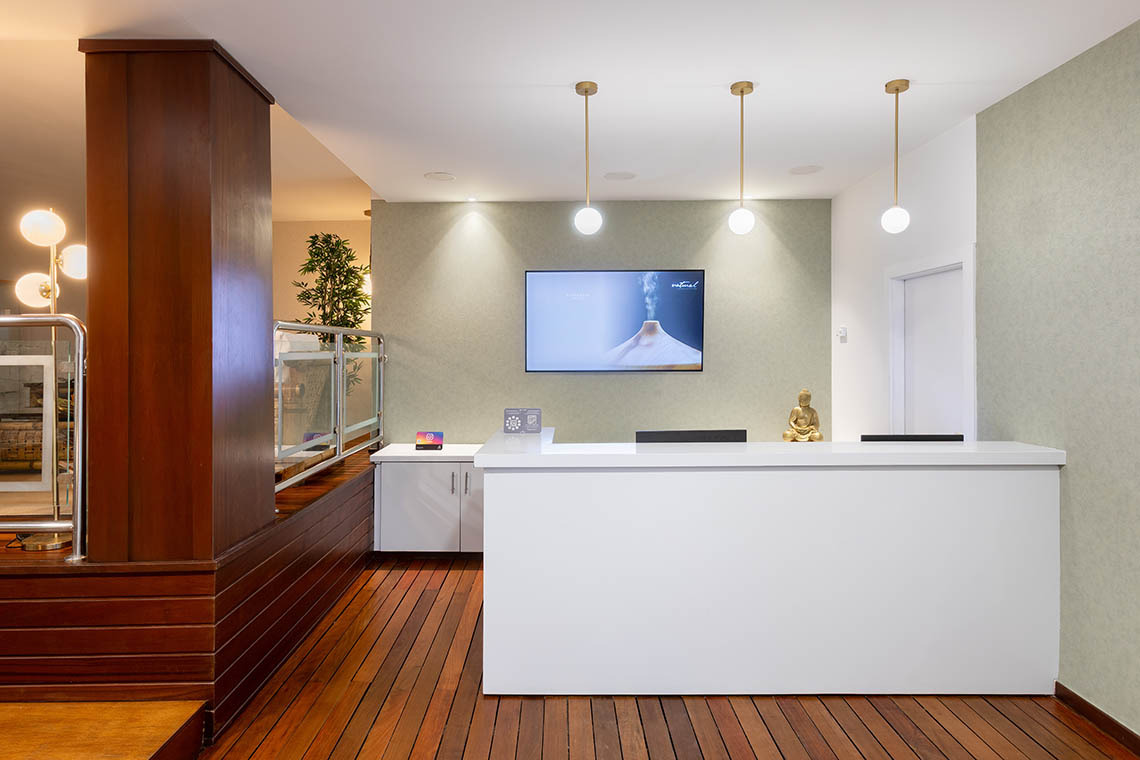 Recepción moderna Alexandre Corporativa: TV, lámparas doradas, escritorio blanco, estatuilla de Buda y suelo de madera.