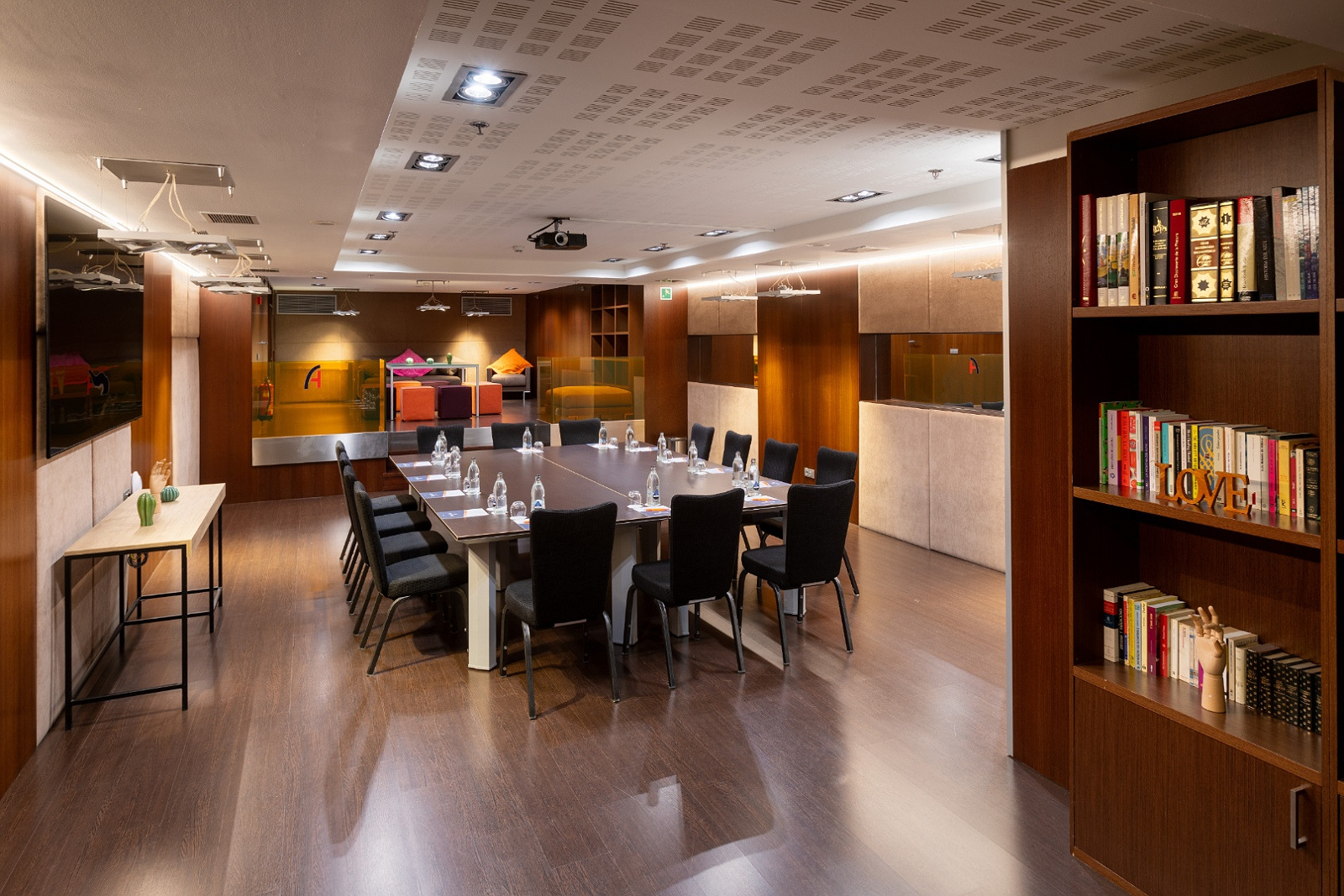 Salle de conférence Alexandre Corporativa moderne, avec table, chaises, projecteur et étagères remplies de livres.