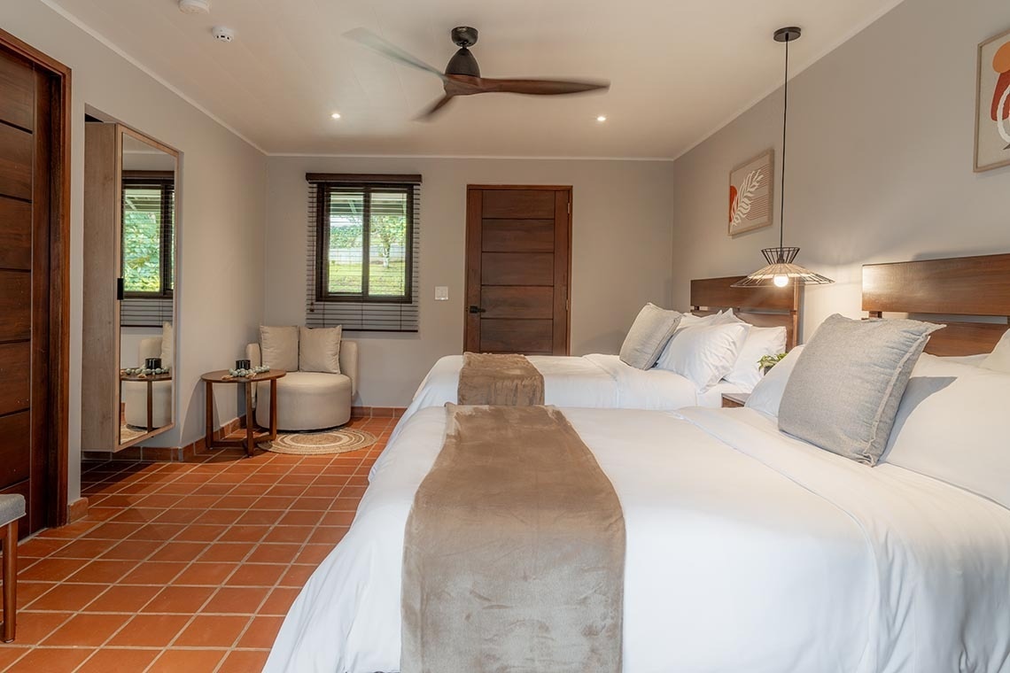 Un luminoso dormitorio de hotel presenta dos camas con cabeceros de madera, paredes grises, un ventilador de techo, suelo de baldosas de terracota y una ventana con persianas de madera.