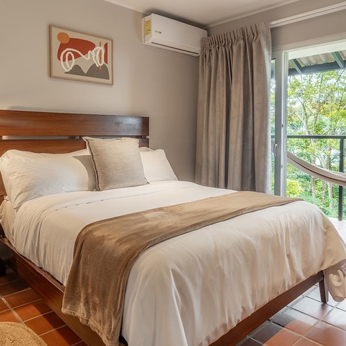 Una habitación moderna con una cama de madera, mesita de noche, decoración abstracta y aire acondicionado, con una puerta de vidrio que se abre a un balcón con hamaca y vista a un paisaje verde.