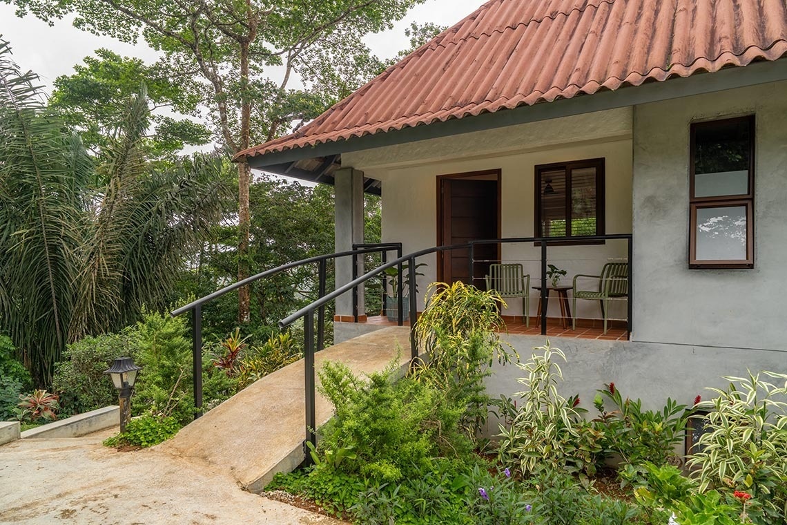Una acogedora casa con tejado rojo y una rampa de acceso está enclavada en medio de un exuberante entorno natural lleno de vegetación tropical.