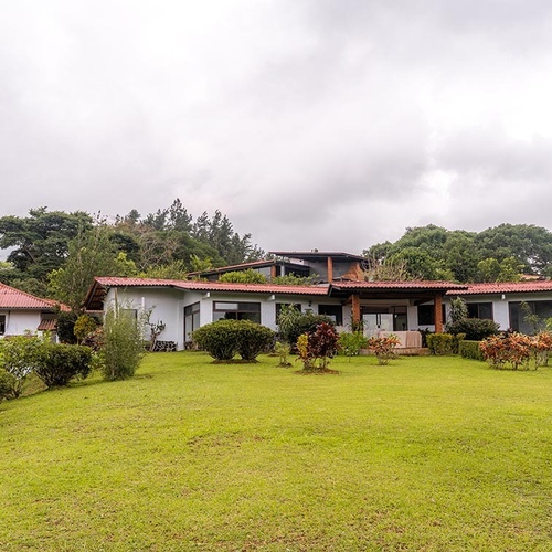 Un conjunto de casas con techos rojos y paredes claras se asienta sobre un césped verde y exuberante, rodeado de abundante vegetación y bajo un cielo nublado.