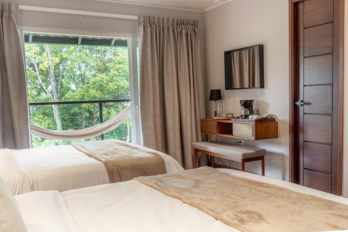 Una habitación de hotel luminosa con dos camas, un balcón con hamaca y vista a la naturaleza, y un rincón de café con un mueble de madera.