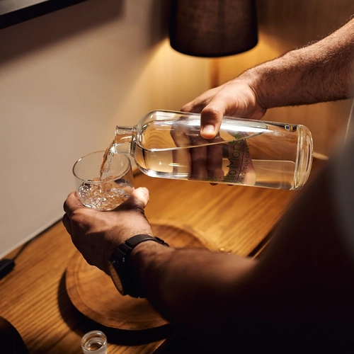 Alto Monte: persona sirviendo agua en un vaso, detalle de la botella con etiqueta y lámpara de noche.