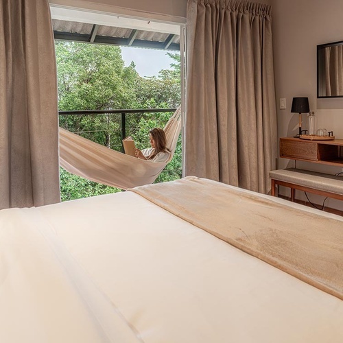 Un dormitorio acogedor y luminoso con una mujer leyendo en una hamaca en el balcón, que da a un entorno natural frondoso, y una estación de café al lado.