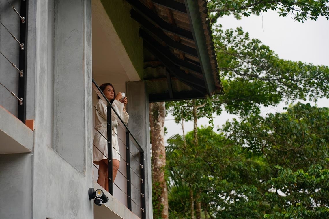 Una mujer en bata de baño sostiene una taza mientras mira el exuberante paisaje de árboles desde un balcón.