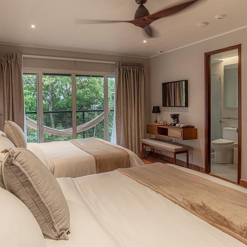 Una moderna habitación de hotel con dos camas, un balcón con hamaca y vistas a la exuberante naturaleza, que incluye un baño visible y un ventilador de techo.