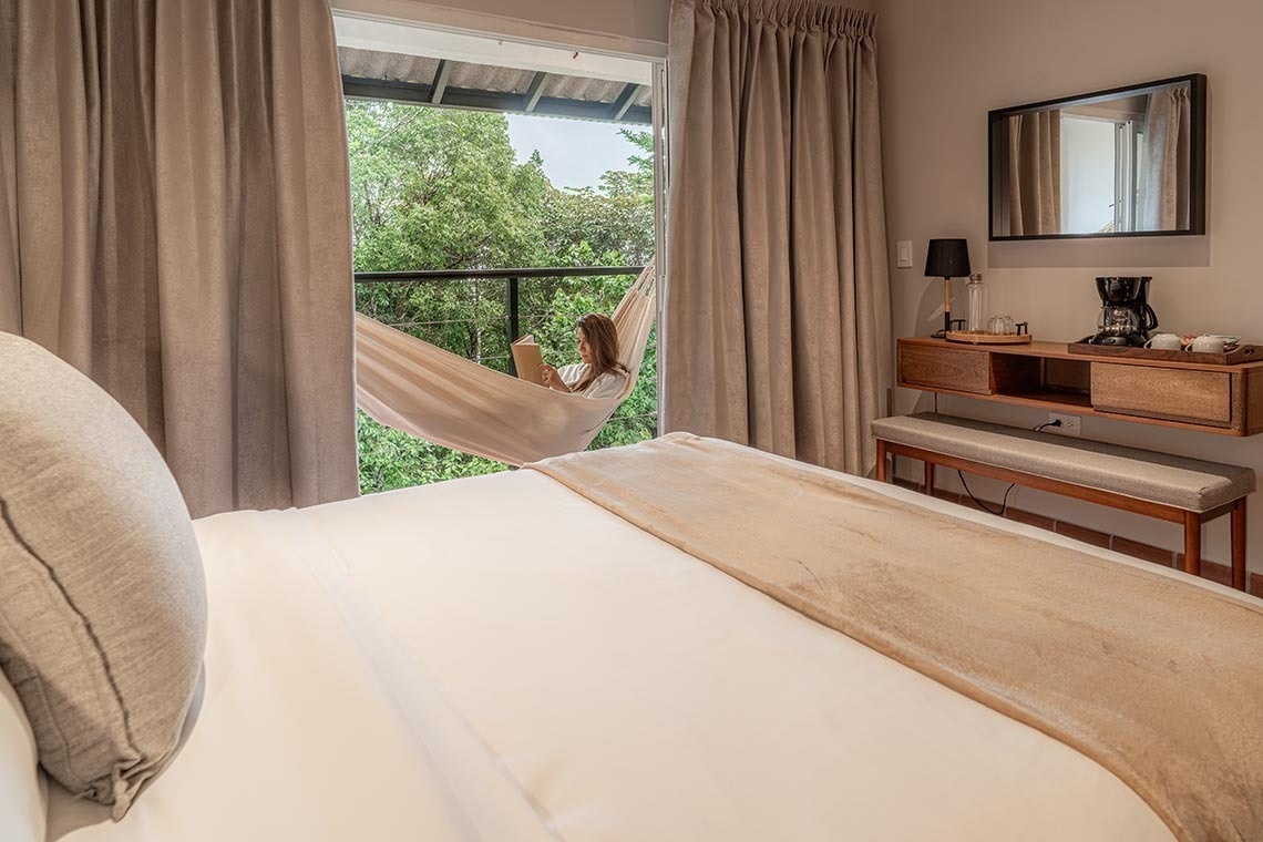 Una mujer lee un libro en una hamaca colgada en el balcón de una habitación, con una cama en primer plano y un paisaje natural de árboles frondosos de fondo.
