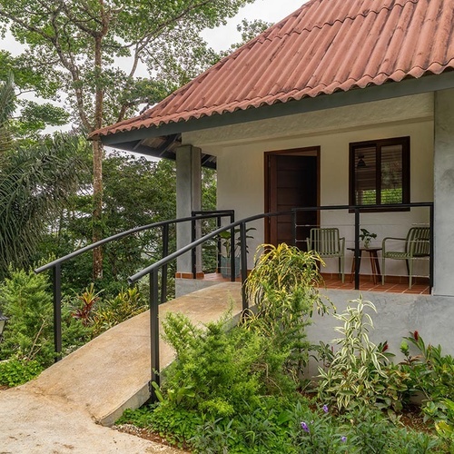 Una pequeña casa con techo de tejas rojas y un porche con rampa de acceso, rodeada de exuberante vegetación tropical.