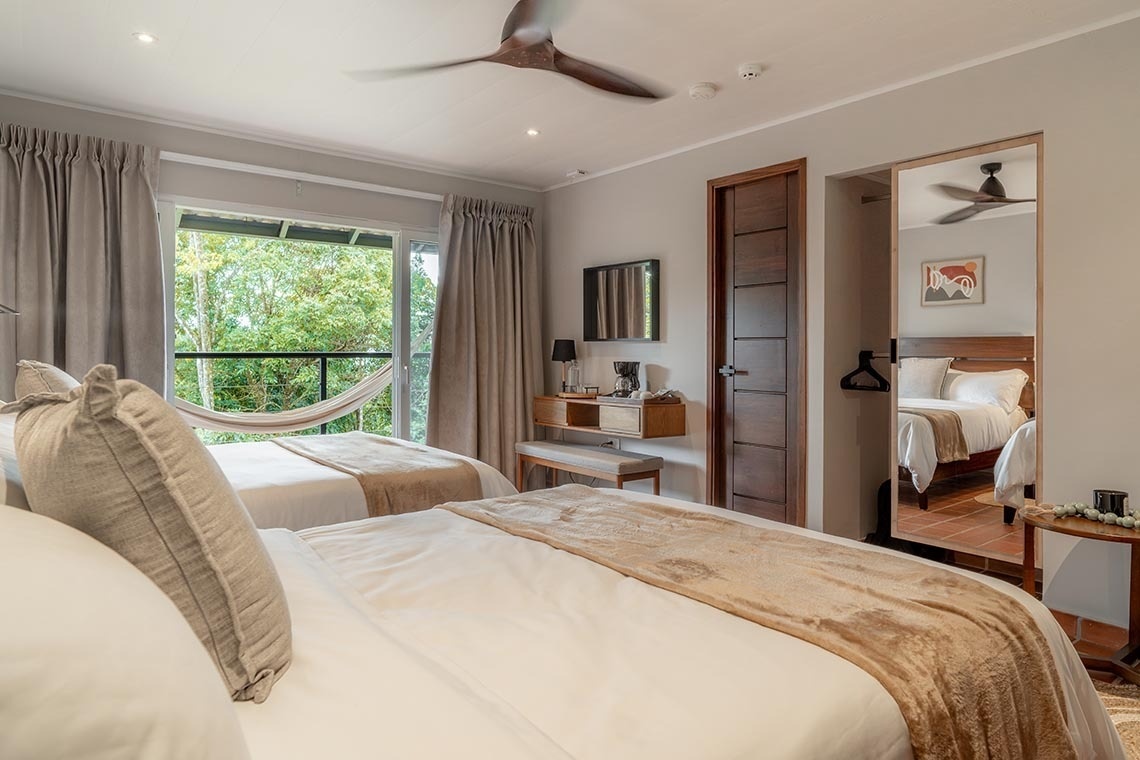 Moderna habitación de hotel con dos camas, un balcón con hamaca, vistas a la exuberante vegetación y un ventilador de techo.