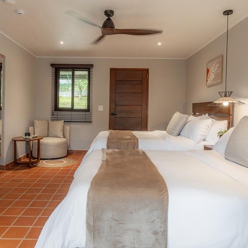 Una acogedora habitación con dos camas, un ventilador de techo, un suelo de baldosas de terracota y vistas a la naturaleza.