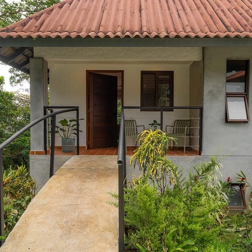 Una casa o cabaña con techo de tejas rojas y un porche con una rampa y barandillas, rodeada de exuberante vegetación tropical.