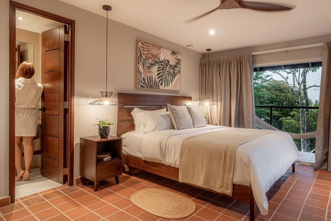 Un dormitorio con suelo de terracota, una cama y mesitas de noche, un gran ventanal con vistas a la vegetación, y una persona en albornoz en la entrada del baño.