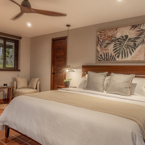 Una habitación acogedora y tropical con una cama grande, muebles de madera, un ventilador de techo, arte de hojas y un suelo de terracota.