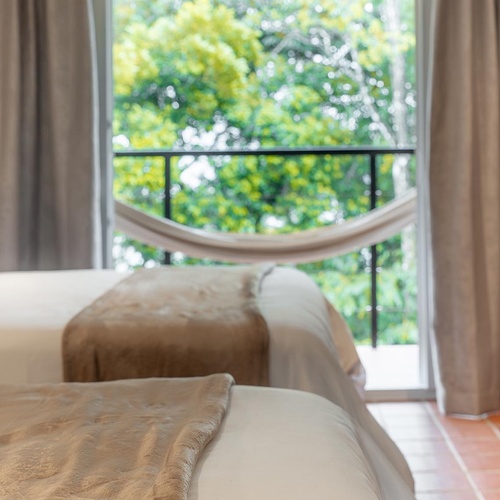 Dos camas con mantas de color beige se ven en primer plano, con una ventana que muestra un balcón con una hamaca y frondosos árboles verdes al fondo.