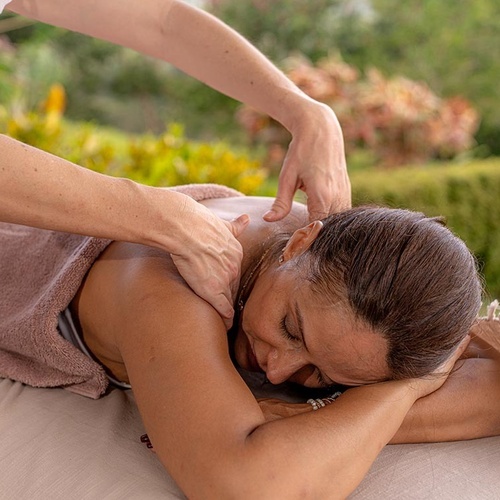 Una mujer recibe un relajante masaje de espalda al aire libre, con los ojos cerrados, mientras unas manos le masajean los hombros y la parte superior de la espalda.