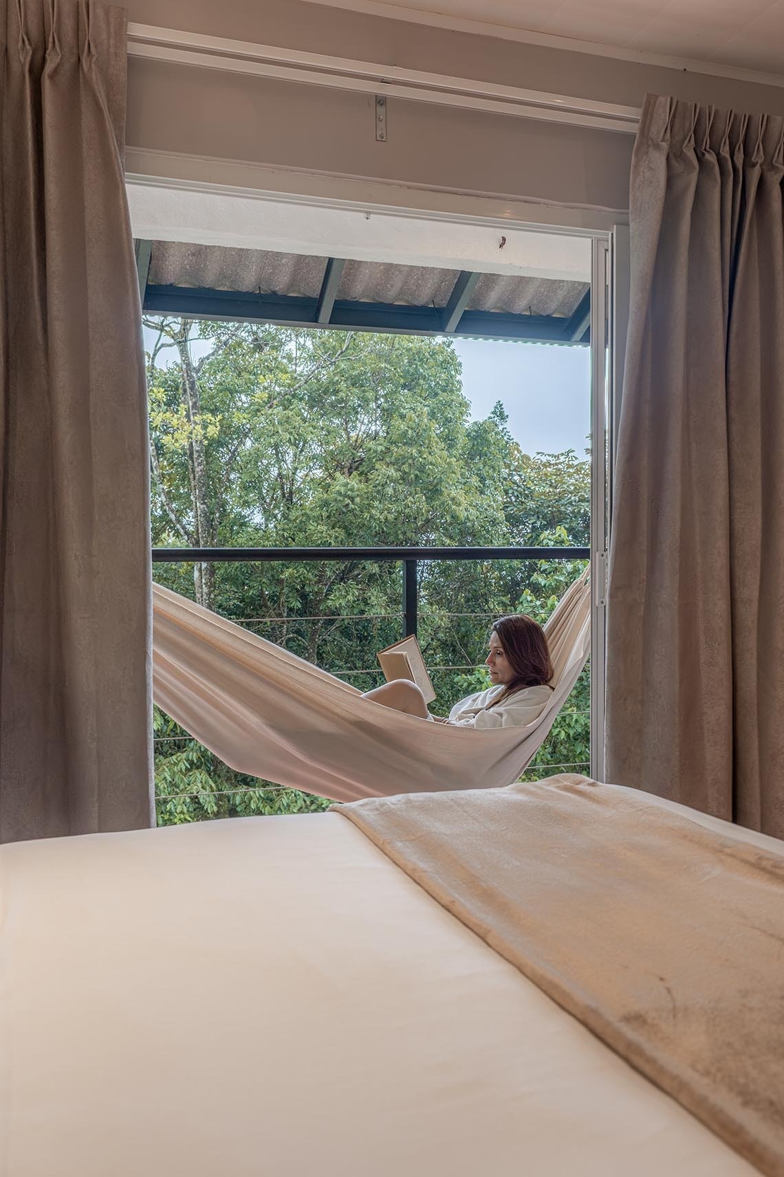 Desde una habitación, una mujer lee un libro en una hamaca en un balcón con vista a un frondoso bosque.