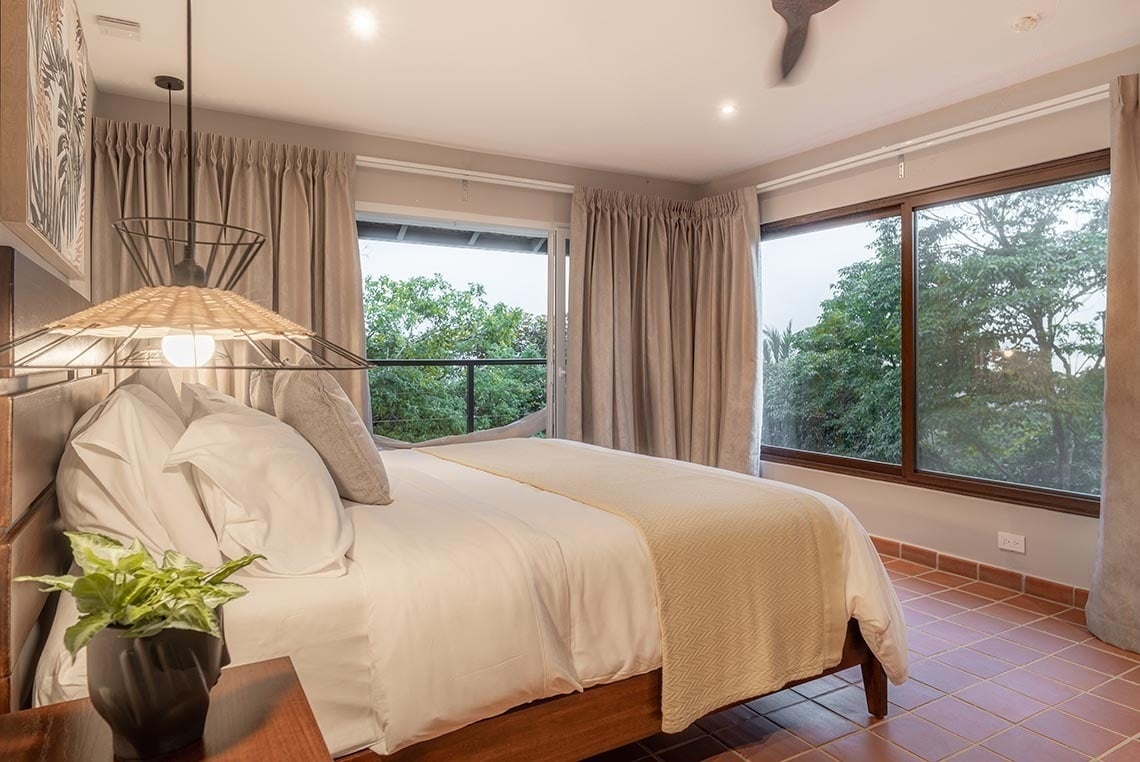 Un dormitorio moderno y luminoso con una cama grande, ventanales que ofrecen vistas a la vegetación exterior, un suelo de baldosas de terracota y una lámpara colgante con pantalla de mimbre.