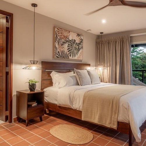 Una habitación de hotel tropical presenta una cama grande, suelo de terracota, una ventana con vista a la vegetación y una persona en el umbral de una puerta.