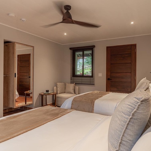 Una habitación moderna con dos camas individuales, puertas de madera oscura, un ventilador de techo y una zona de asientos junto a una ventana.