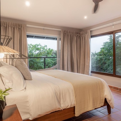 Este es un dormitorio luminoso con una cama bien hecha, una lámpara colgante de mimbre, grandes ventanales que dan a un paisaje verde exuberante y un suelo de baldosas de terracota.