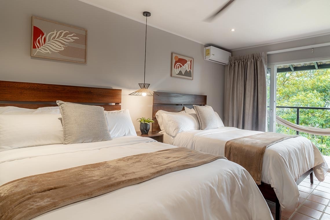 Un dormitorio doble moderno y luminoso con cabeceros de madera, ropa de cama blanca y detalles beige, arte en la pared y un balcón con hamaca que ofrece vistas a la vegetación.