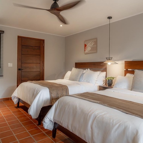 Un luminoso dormitorio cuenta con dos camas con cabeceros de madera, ropa de cama blanca, suelo de baldosas de terracota, un ventilador de techo y una ventana con persianas.