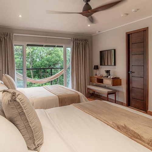 Una espaciosa habitación de hotel con dos camas, un ventilador de techo, muebles de madera y un balcón con hamaca que ofrece vistas a un frondoso bosque.