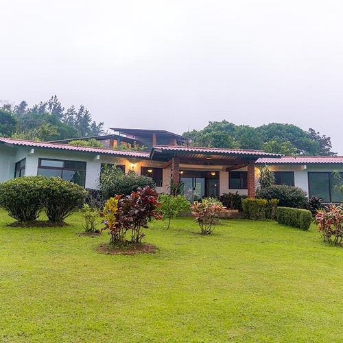 Una casa moderna de un solo piso con techo de tejas rojas se erige en medio de un extenso césped verde y exuberante vegetación, bajo un cielo nublado.