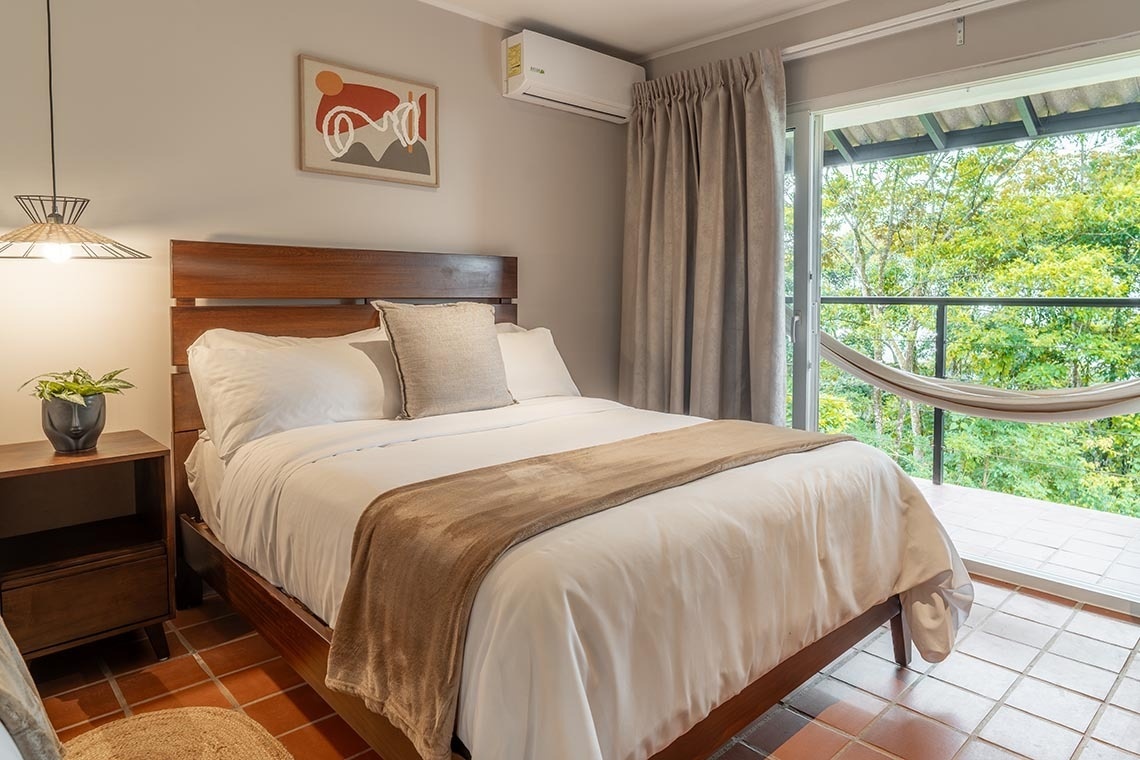 Un acogedor dormitorio cuenta con una cama de madera y ropa de cama clara, decorado con un cuadro moderno, y se abre a un balcón con hamaca que ofrece una exuberante vista verde.