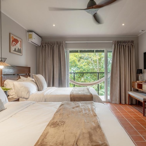 Una luminosa habitación de hotel cuenta con dos camas, un ventilador de techo, aire acondicionado y un balcón con hamaca que ofrece vistas a una exuberante vegetación.
