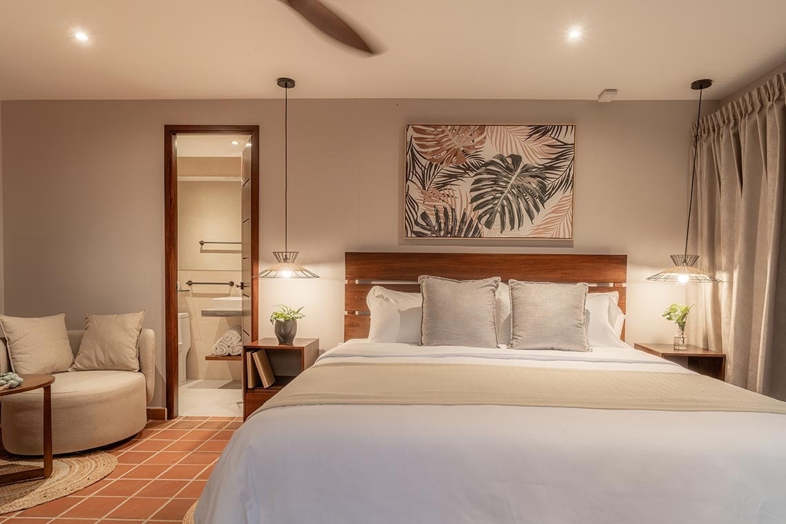 Un dormitorio moderno y luminoso con una cama grande, cabecero de madera, decoración tropical, zona de estar y un baño visible, todo sobre un suelo de terracota.