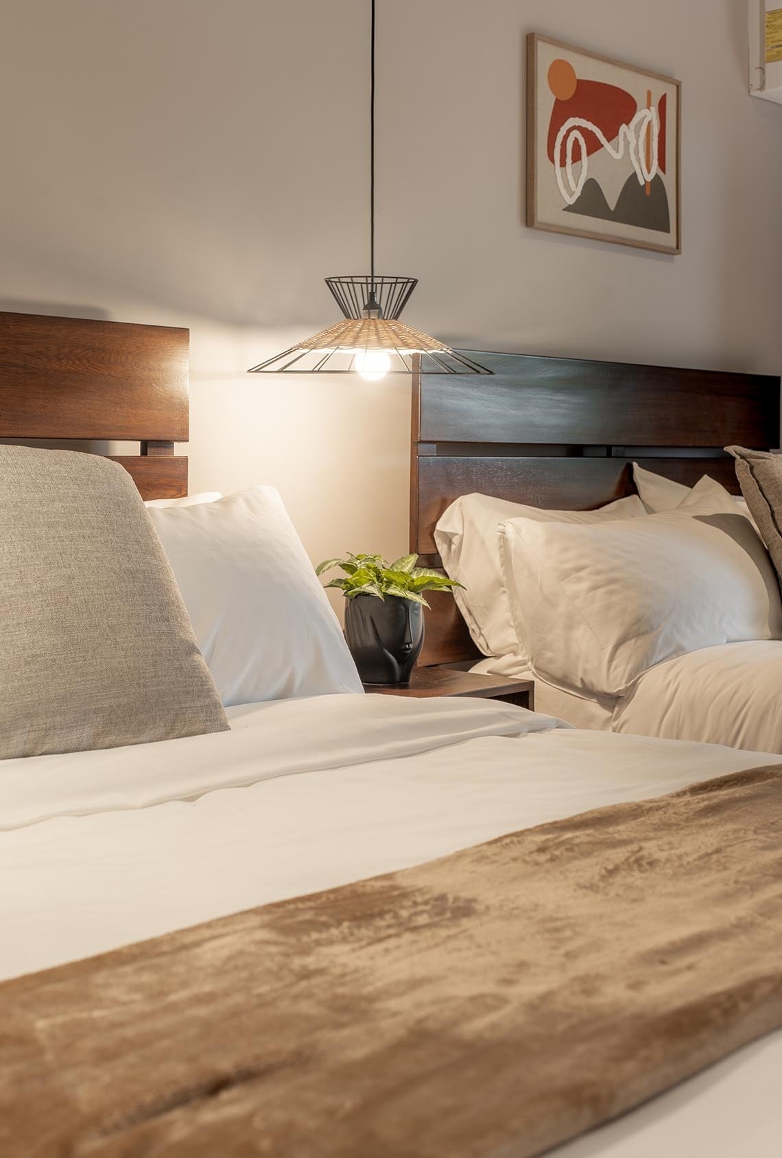 Un acogedor dormitorio presenta dos camas con cabeceros de madera oscura, una lámpara colgante moderna y una obra de arte abstracta enmarcada en la pared.